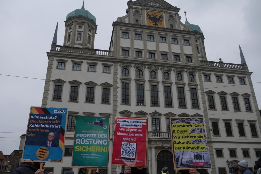 Ein Foto von Wahlschildern im Design von CDU, Grünen, SPD und FDP, die vor dem Augsburger Rathaus in die Höhe gehalten werden. Auf dem Schild im Design der CDU steht: "Klimaschutz? Blockieren wir! Kohleindustrie? Profitieren wir! Klima juckt uns nicht". Auf dem Schild im Design der Grünen steht: "Klimaschutz ist out! Profite der Rüstung sichern! Ein Bündnis. 100 Milliarden für die Bundeswehr". Auf dem Schild im Design der SPD steht: "Ausstieg aus fossilen Brennstoffen? Wir zögern es hinaus, bis es zu spät ist!". Auf dem Plakat im Design der FDP steht: "Atomkraft, Gas und Diesel. Wir sichern die Zukunft. Der Klimakiller."