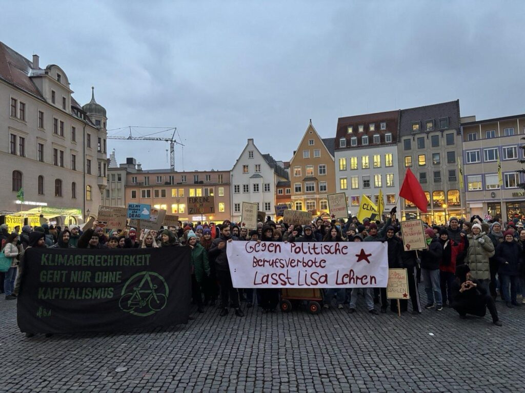 Viele Menschen stehen auf dem Augsburger Rathausplatz und gucken in die Kamera. Sie halten Demoschilder mit Sprüchen für Klimagerechtigkeit und vorne links halten Menschen ein schwarzes Banner mit der grünen Aufschrift "Klimagerechtigkeit geht nur ohne Kapitalismus" neben einem grünen Anarchie-A, in dessen Design ein Fahrrad integriert ist. Rechts vorne halten Menschen ein großes Papier mit der roten Aufschrift "Gegen politische Berufsverbote. Lasst Lisa lehren" und daneben einem roten Stern.