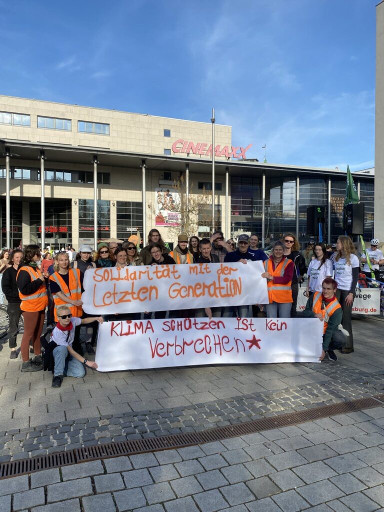 Das Bild zeigt einige Menschen, die vor der City Galerie in Augsburg stehen und gemeinsam zwei große Banner aus Papier halten. Auf dem einen Banner steht in orangefarbener Schrift: "Solidarität mit der Letzten Generation". Auf dem Banner, das darunter gehalten wird, steht in roter Schrift: "Klima schützen ist kein Verbrechen" und daneben ist ein roter Stern aufgemalt.